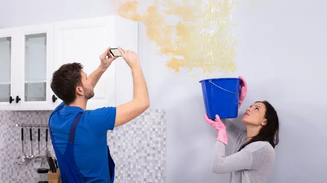 Man photographing a water-stained ceiling with his phone while a woman holds a blue bucket under the drip
